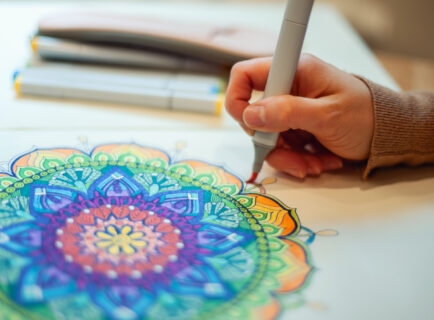 Young woman colouring mandala with markers and white rosary on table with cup of coffee at home Young woman colouring mandala with markers and white rosary on table with cup of coffee near flower vase at home. Art therapy and meditation concept. Hobby and leisure during quarantine. Wellbeing.